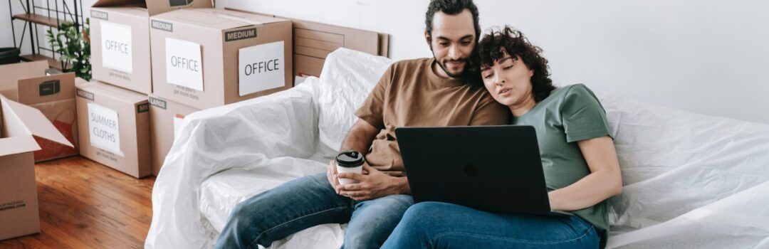 couple sitting on couch using laptop with moving boxes near by